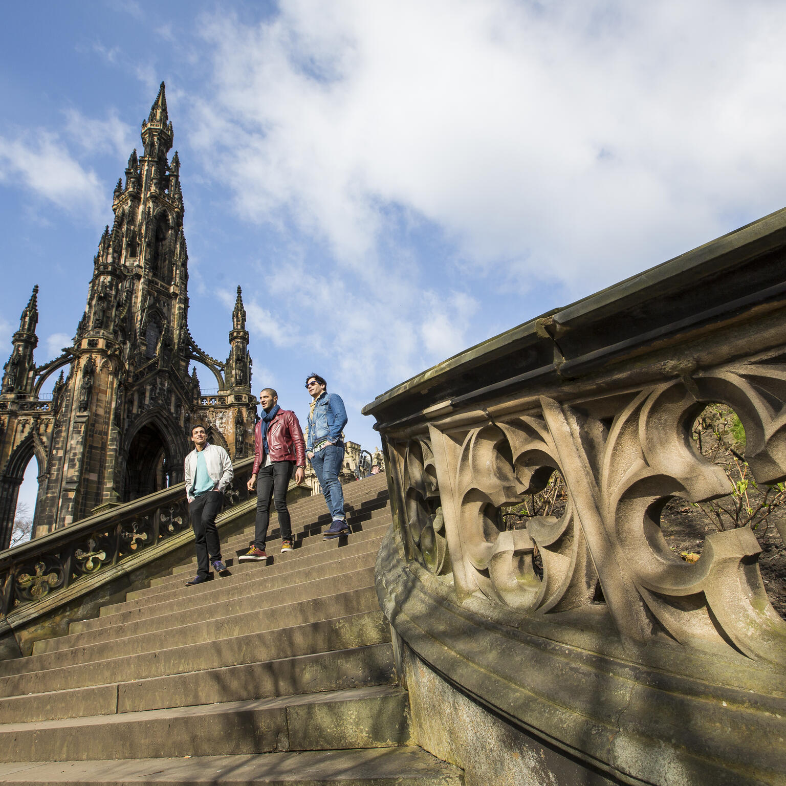 Three people walking down stone steps near the ornate Scott Monument under a partly cloudy blue sky in Edinburgh.