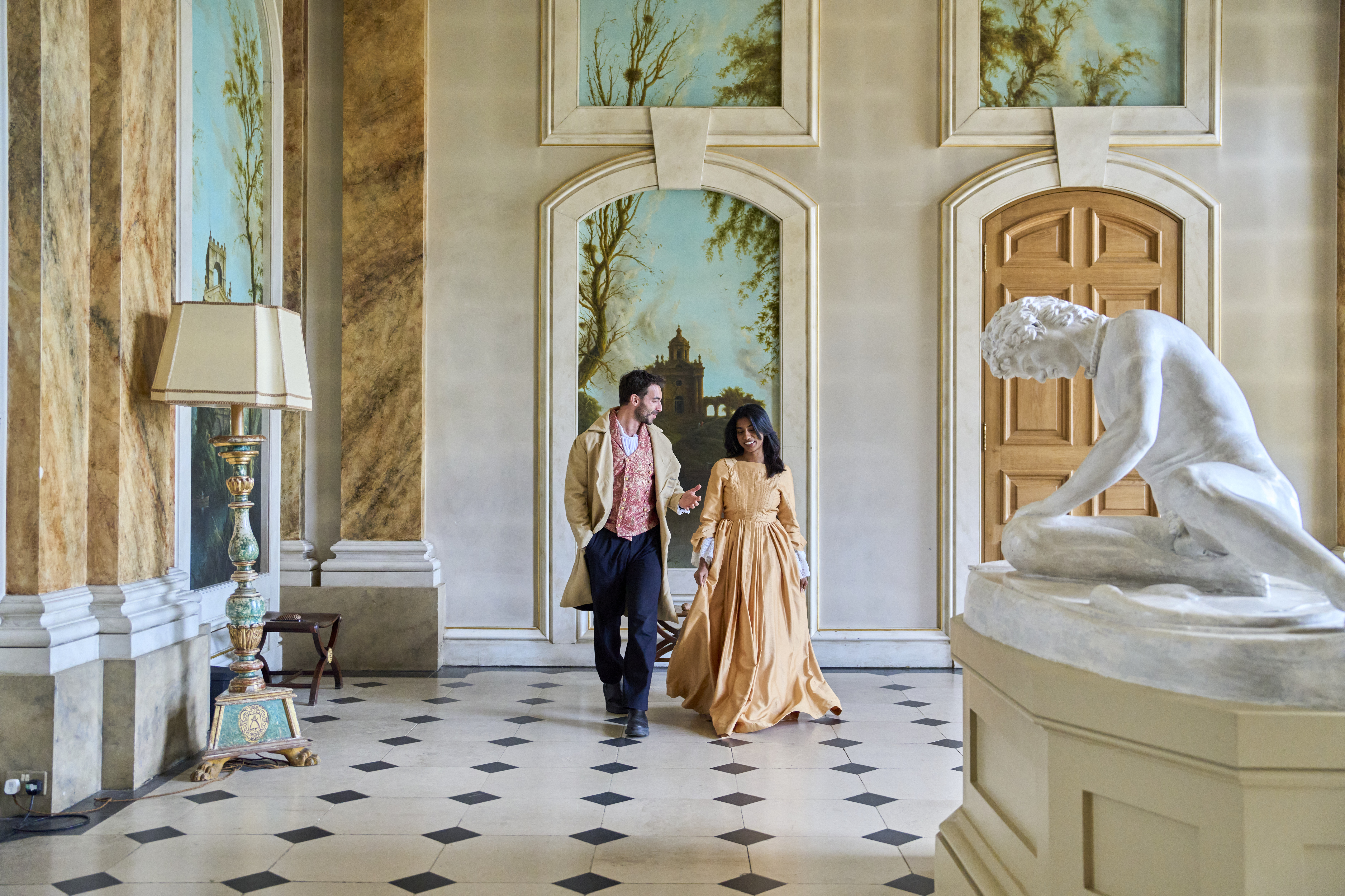 Elegant hall with classical decor, statue, ornate lamp, and two people in period attire walking and talking. Arched murals and wooden doors in the background.