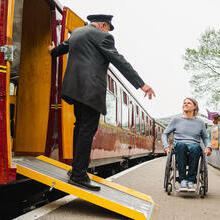 Guard stands on a ramps and leans out of heritage train carriage greeting woman and man using a wheelchair