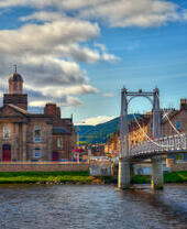A suspension bridge with people walking across spanning a wide river with houses on the other side