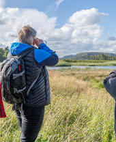 Three people birdwatching in a grassy field by a lake, under a partly cloudy sky, with mountains in the distance.