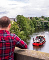 Dos personas de pie en un puente contemplando un barco turístico que navega por un río en Durham