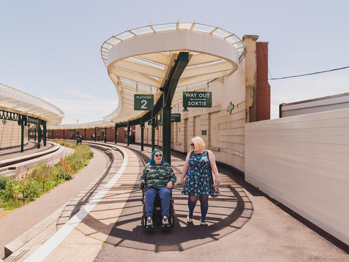 Una mujer camina con otra mujer que está sentada en una silla de ruedas en una antigua estación de tren.