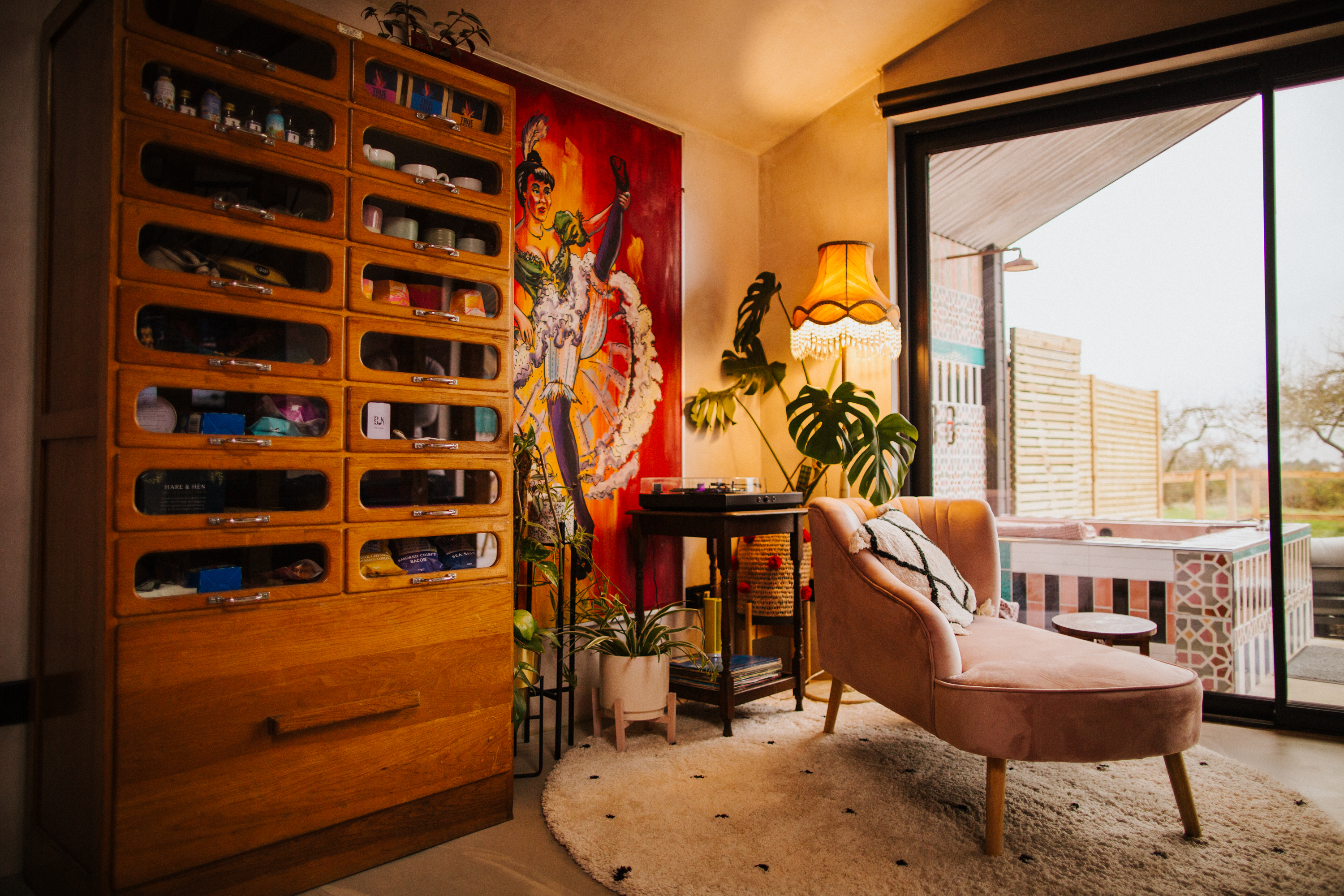 A pale pink chaise lounge on a rug lookng out of floor to ceiling windows. A tall wooden dresser to the left and vibrant artwork on the walls