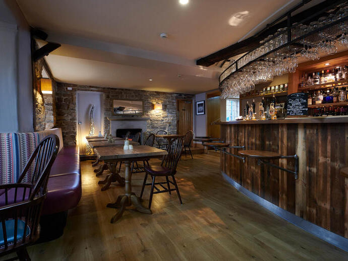 Interior of a pub with the bar area, showing bench seating and dining room tables and chairs, exposed stone walls and a stone fireplace in background