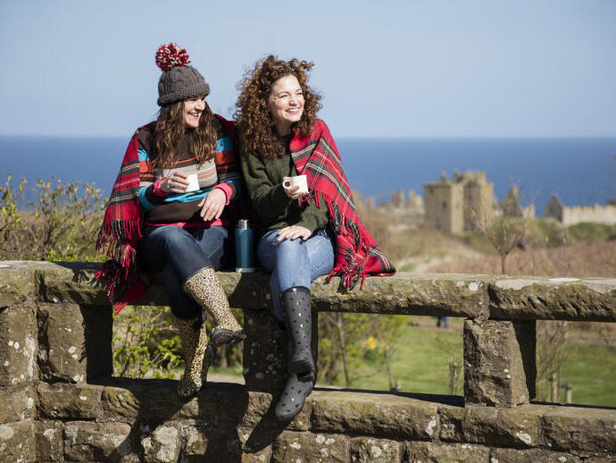 Two women with hot drinks, sitting on a stone wall near a castle