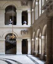 Grand hall with classical architectural features, arched windows, marble floors, ornate chandelier, and sculptures on display.