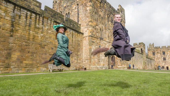 Two wizarding professors demonstrating flying on a broom outside a large castle.