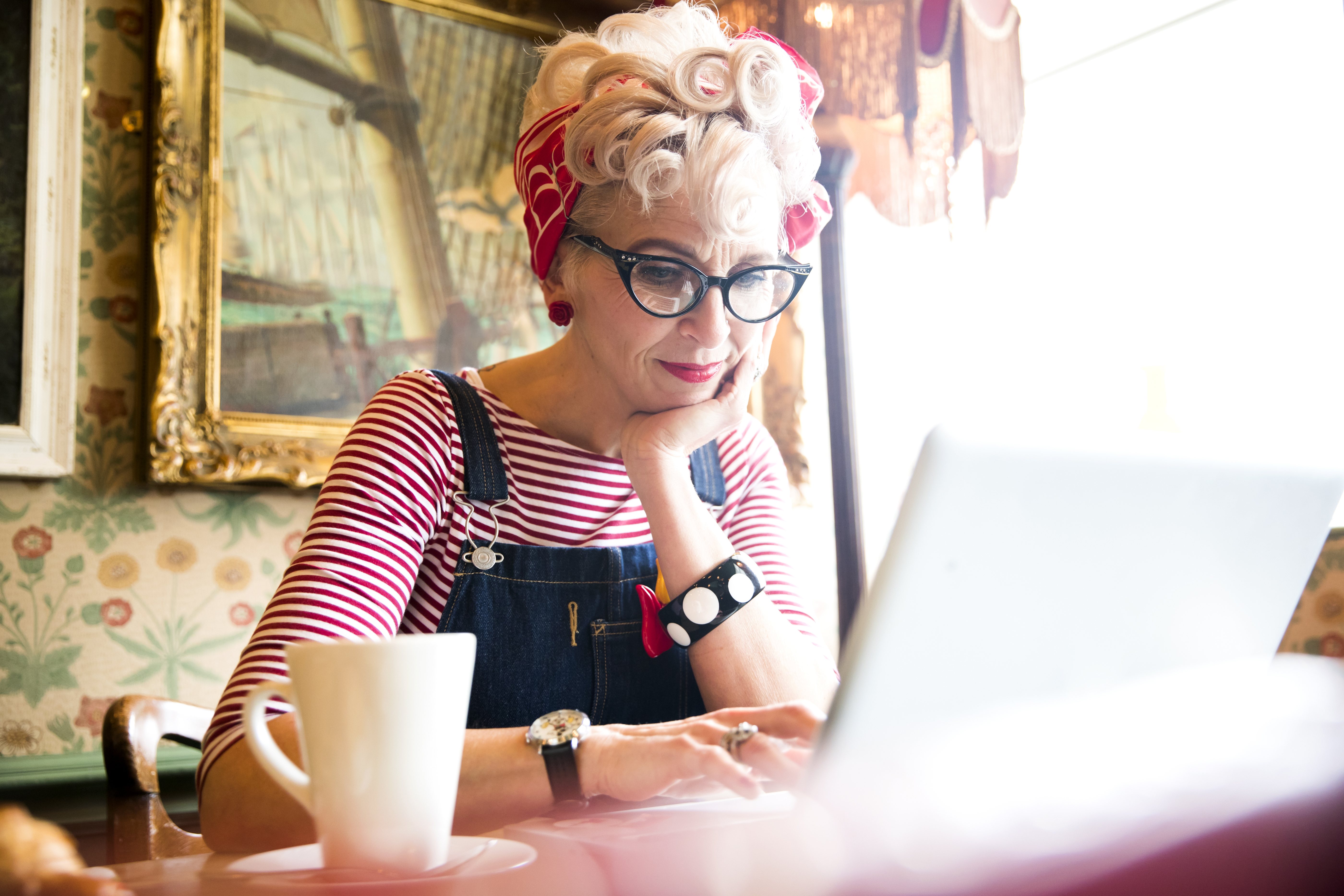 Close up of a blond woman wearing a stripy top and red head scarf, having a coffee and looking down at a lap top screen, smiling.