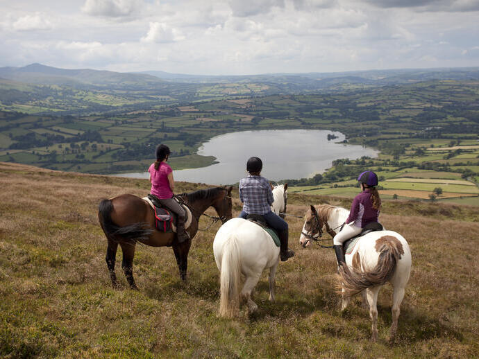 Eine Gruppe von Reitern auf einem Hügel mit Blick auf Ackerland und einen See.