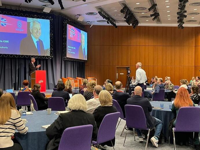 El presidente de la BTA, Nick de Bois CBE, de pie detrás de un podio en el escenario, se dirige a una sala con 80 delegados en la Conferencia de la Asociación de Eventos Empresariales de VisitBritain.