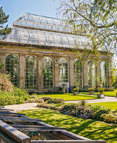 Large glasshouse with arched windows in a botanical garden, surrounded by green lawns, trees, and landscaped flower beds on a sunny day.