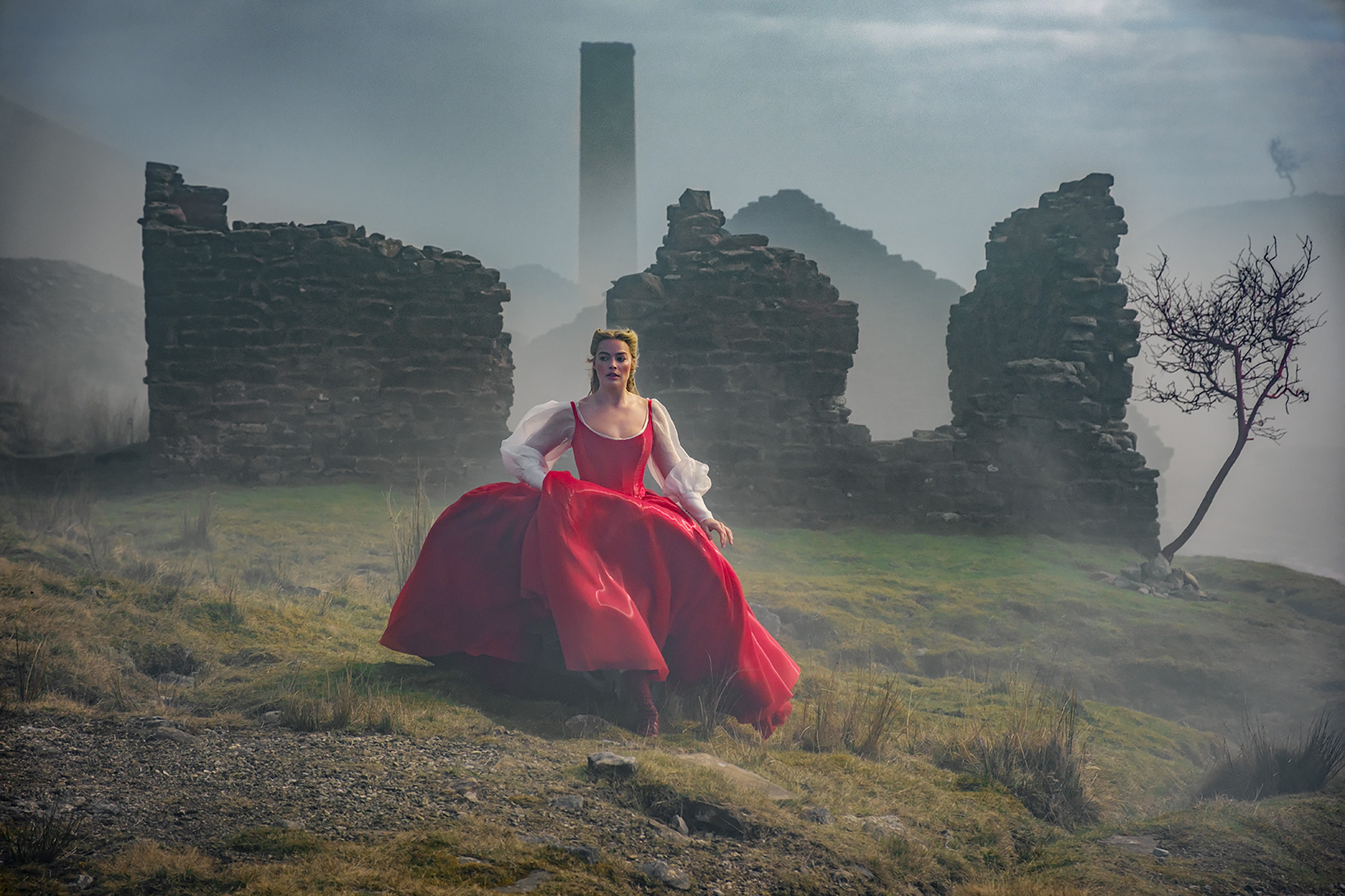 A person in a vibrant red dress stands on grass near ruins and a tree, with mist and hills in the background, evoking a dramatic scene.