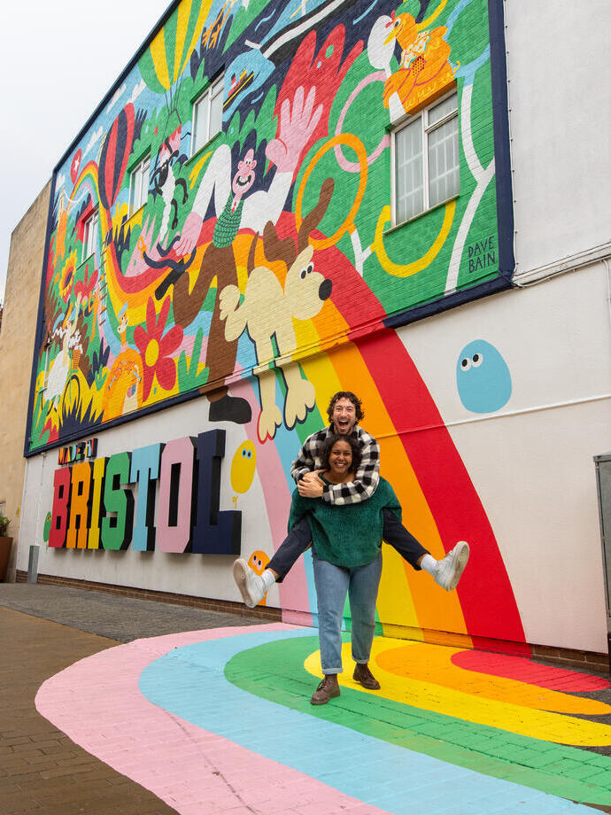  Two people playfully smiling and posing in front of a colourful mural.
