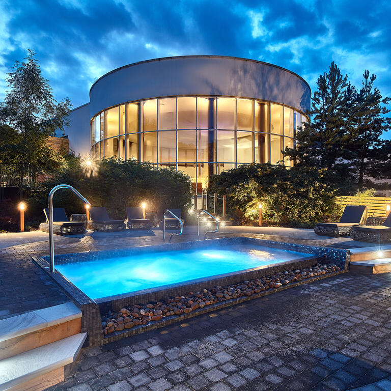 The outdoor spa area at dusk with a lit outdoor spa pool surrounded by loungers and the glass fronted circular spa building in the background. 