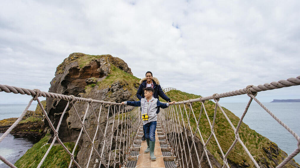 Two people crossing a rope bridge over cliffs with the sea and rocky, grassy landscape in the background.