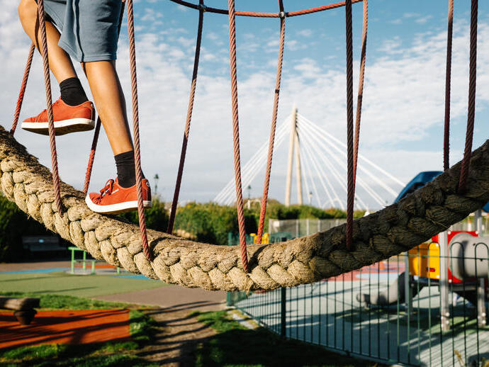 A child wearing red shoes and shorts uses a rope play structure at a playground, with a cable-stayed bridge in the background.