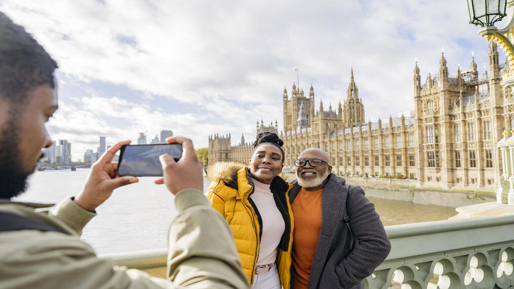 A man photographing mature couple with a large river and iconic buildings in the background.