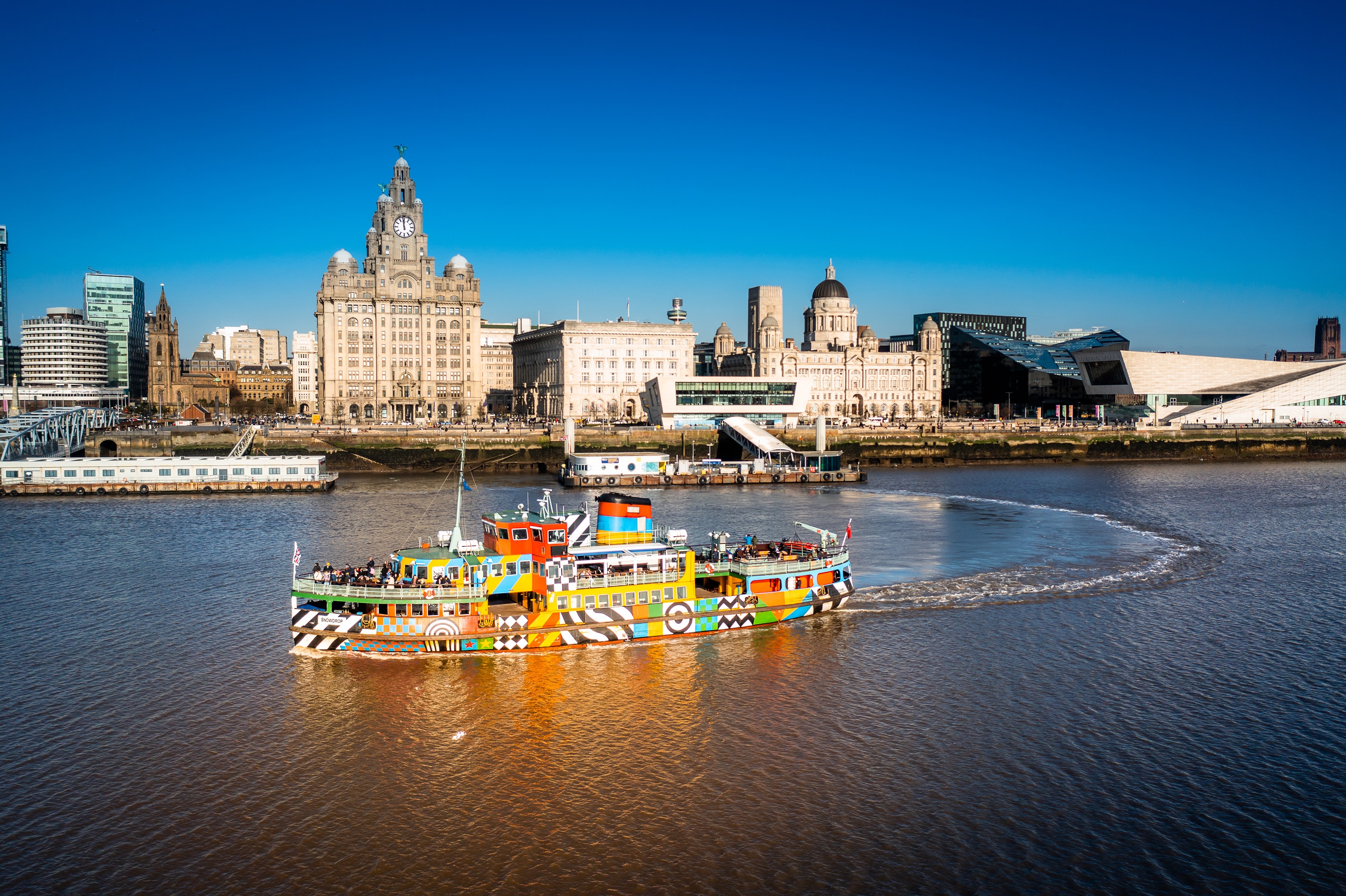 Colorful ferry on the River Mersey with Liverpool waterfront, historic buildings, and modern architecture under a clear blue sky.