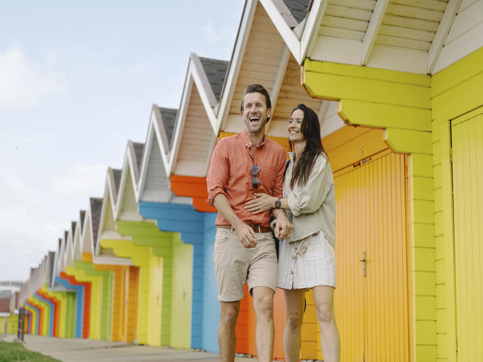 A man and woman laughing outside colourful beach huts