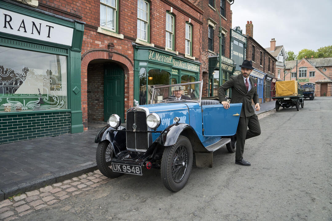 Alte Backsteinstraße mit Geschäften und einem blauen Oldtimer, geparkt am Straßenrand, ein Mann im Anzug steht an der Tür.