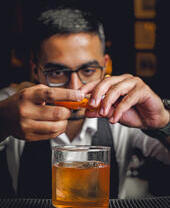 Hombre preparando un cóctel, exprimiendo cáscara de naranja sobre un vaso con bebida alcohólica y hielo en la barra.