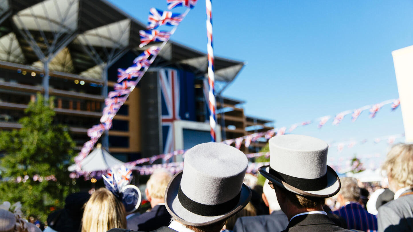 Two men in formal dress with top hats in a crowd with a grandstand and bunting in the distance