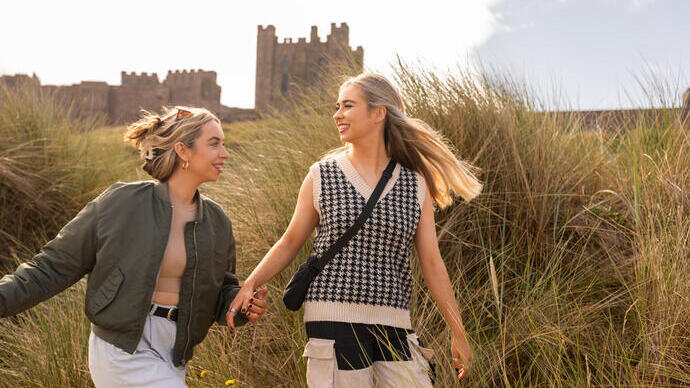 Two women holding hands and walking in the sand dunes near by the sea with a castle in the background