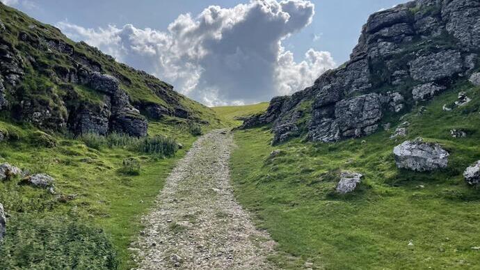 Rocky footpath with rocky valley walls starting to drop. Low-level large clouds are in the background.