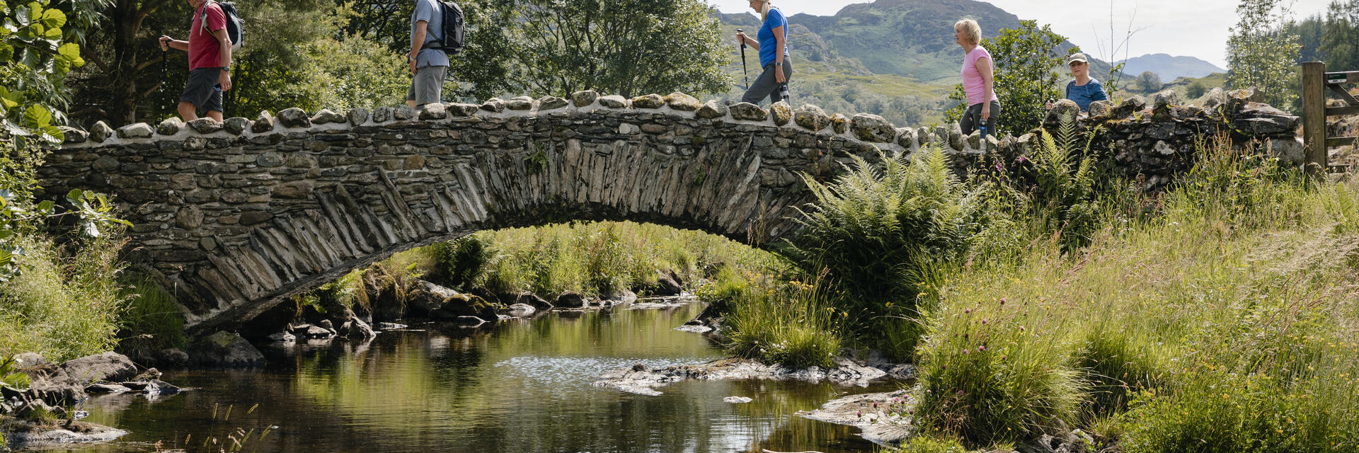 Un groupe de personnes marche sur un pont en pierre au-dessus d'une rivière, entouré de verdure et de collines sous un ciel ensoleillé.