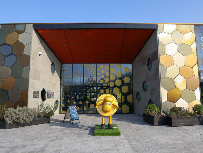 Modern building with hexagonal patterns, glass entrance, and sheep statue at the front, under a clear blue sky.
