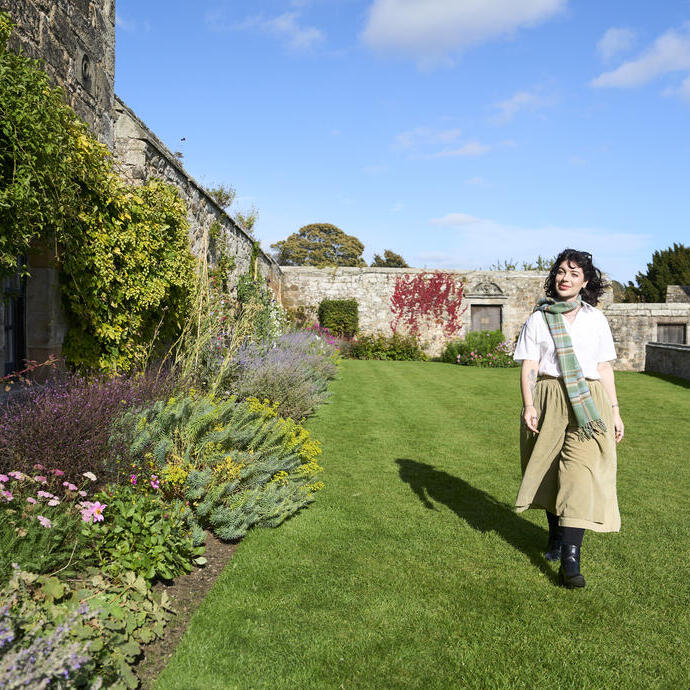 Frau geht im Garten neben einer Steinmauer, umgeben von Blumen und Pflanzen, unter blauem Himmel.