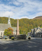 Ein kleiner Dorfplatz mit einer Kirche, einigen Steinhäusern und einem roten Denkmal in der Mitte.