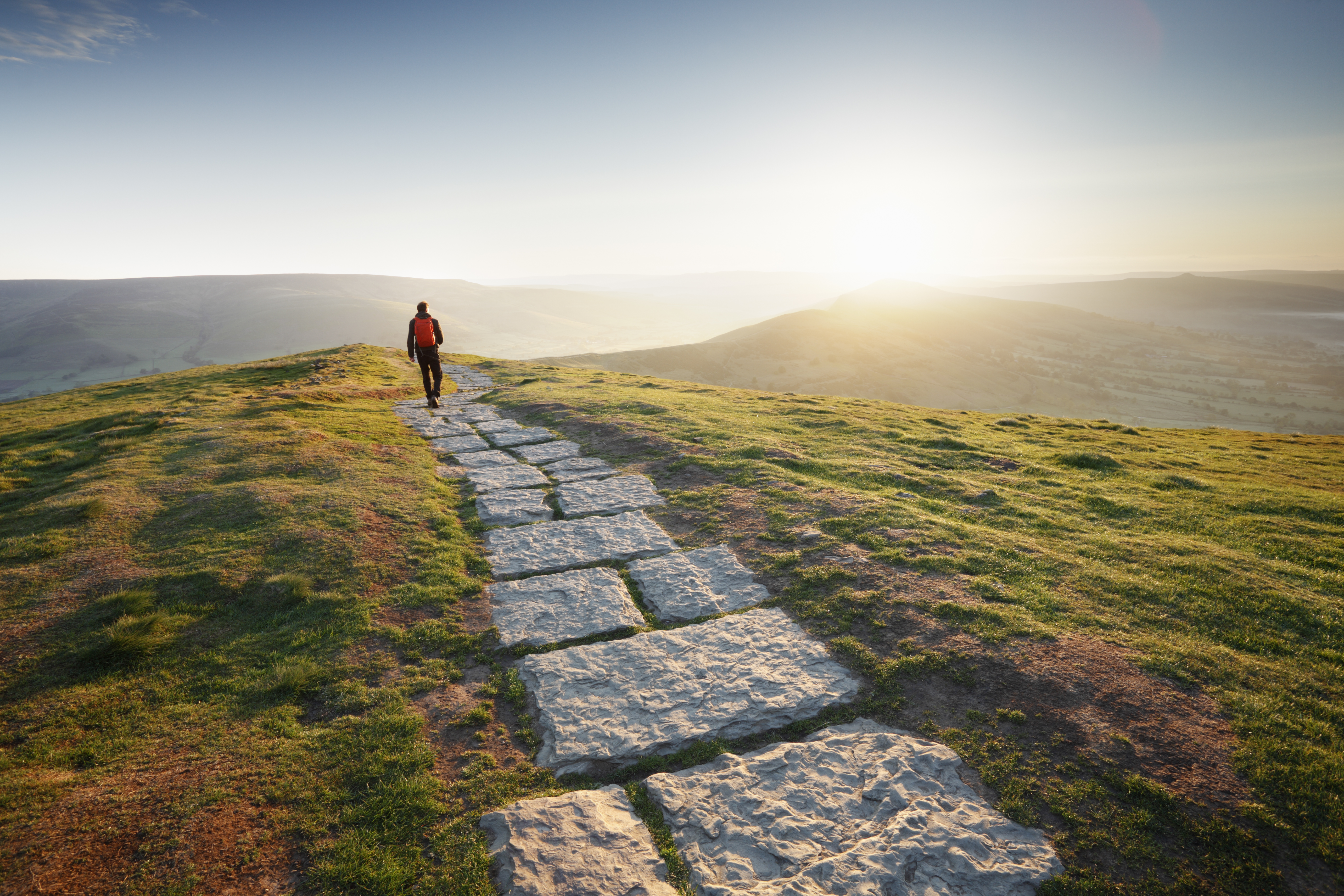 Man in red jacket walking away into the sun at the top of a mountain path