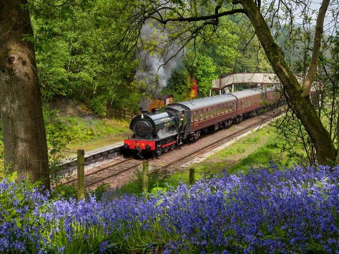 A steam train with lots of steam coming out, pulling at least three vintage carriages, gets ready to depart a small traditional railway station.