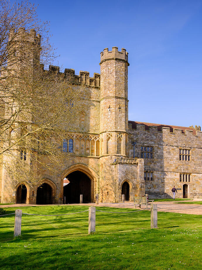 Historic stone building with towers, arched entrances, manicured lawn, trees, and a clear blue sky in the background.