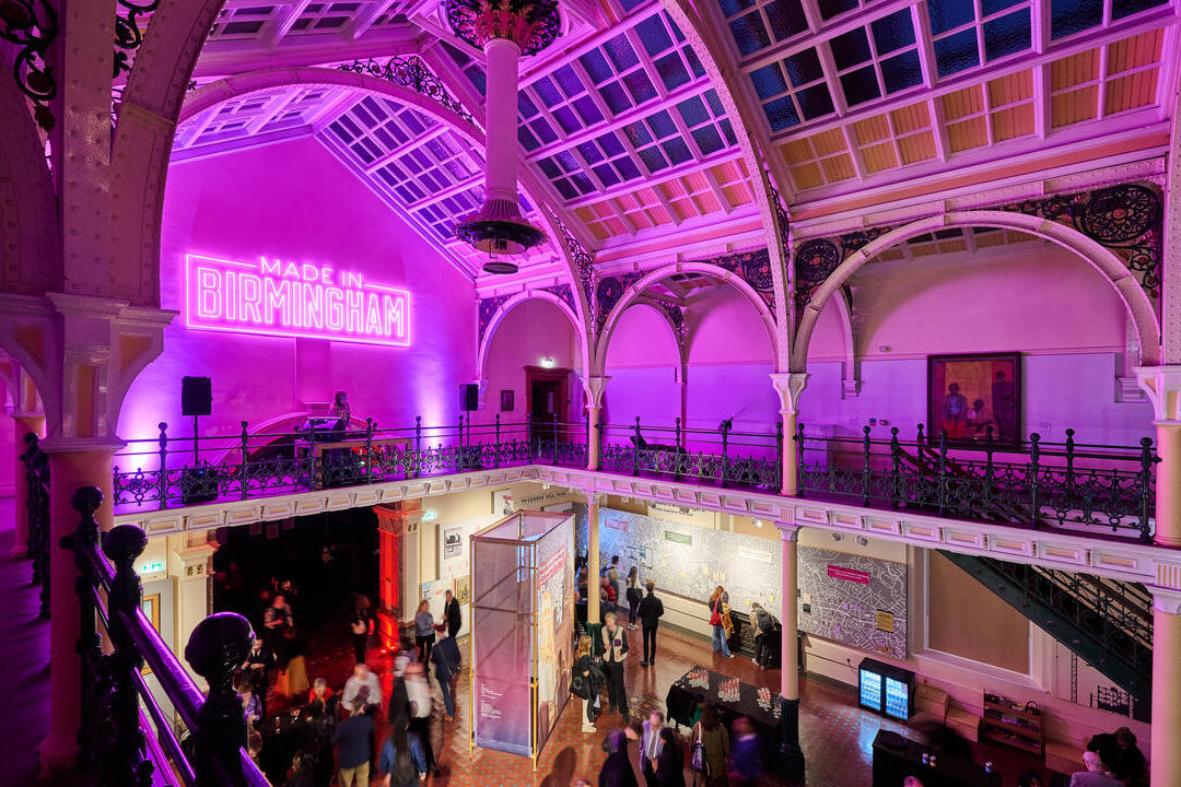 Interior of a grand hall with ornate arches, stained glass ceiling, neon 'Made in Birmingham' sign, and people viewing exhibits below.