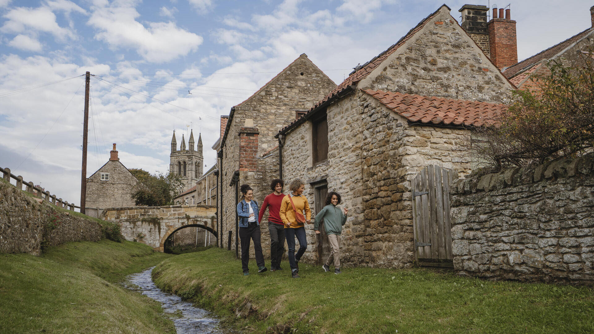 Four people walk beside a stone cottage and stream in a historic village, with a church tower and arched bridge in the background under a partly cloudy sky.