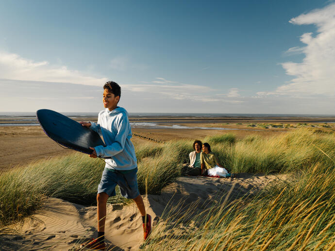 Boy with a surfboard walks on sandy dunes, while two people relax in long grass beside a wide, empty beach under a bright blue sky.