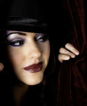 A performer wearing a hat and in full make up peaking out from behind a curtain