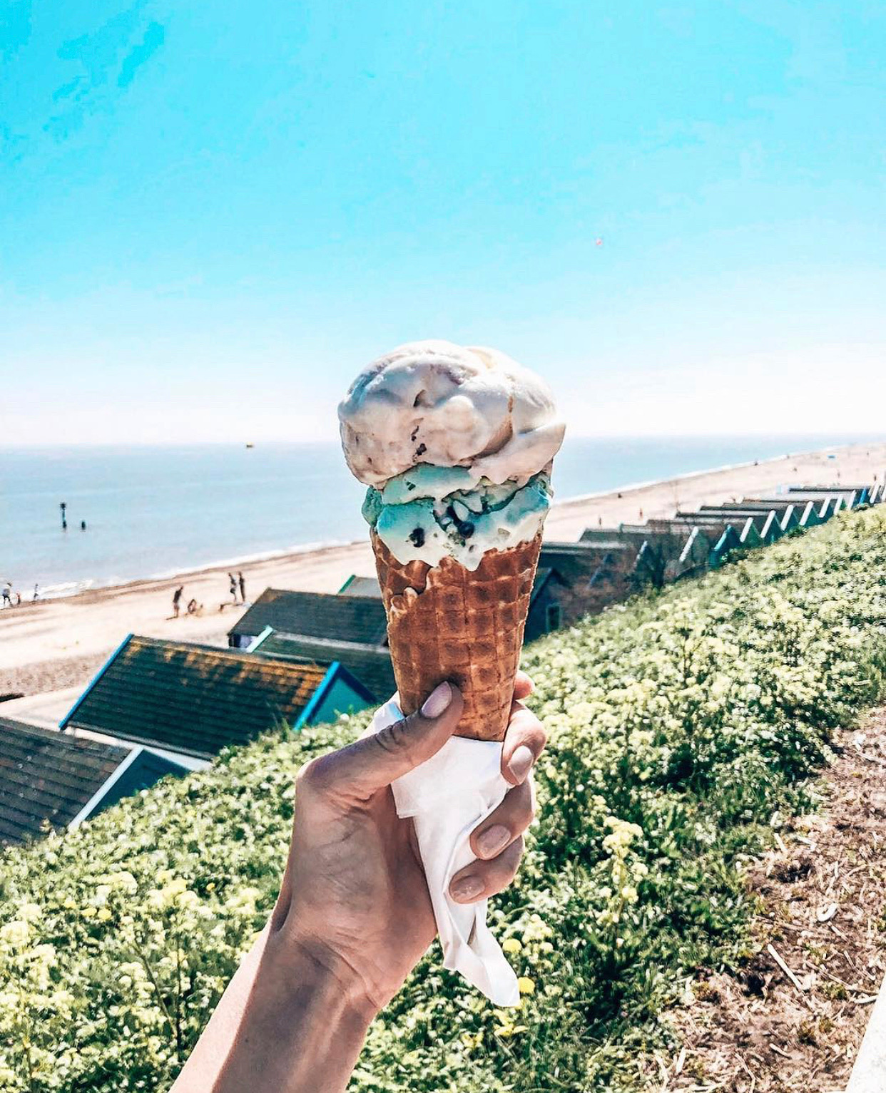 An ice cream cone being held in front of a beach, sea and beach huts