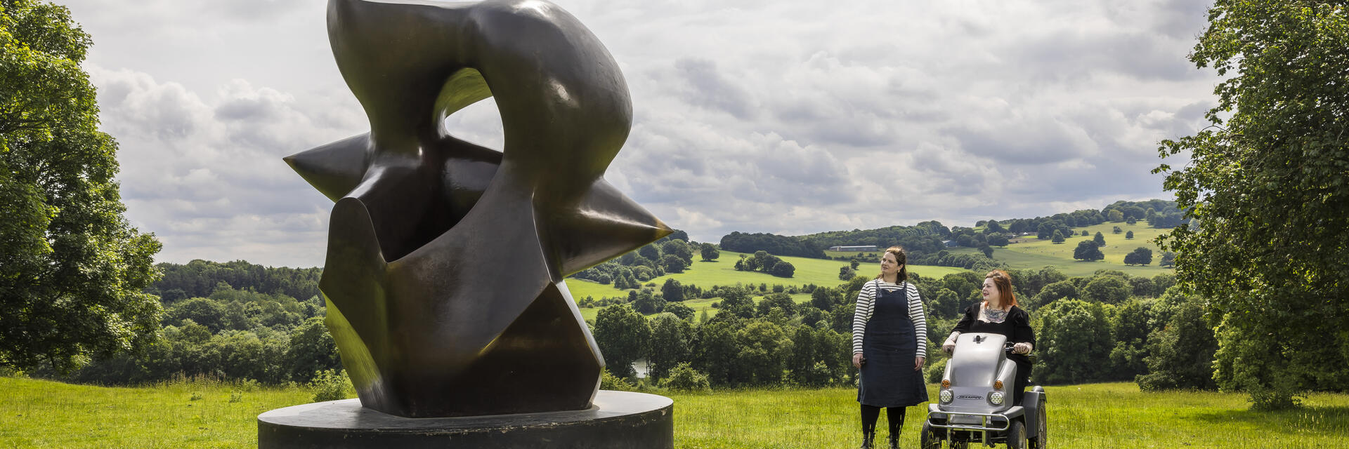 Two women look at a large sculpture set in green gardens with landscape beyond