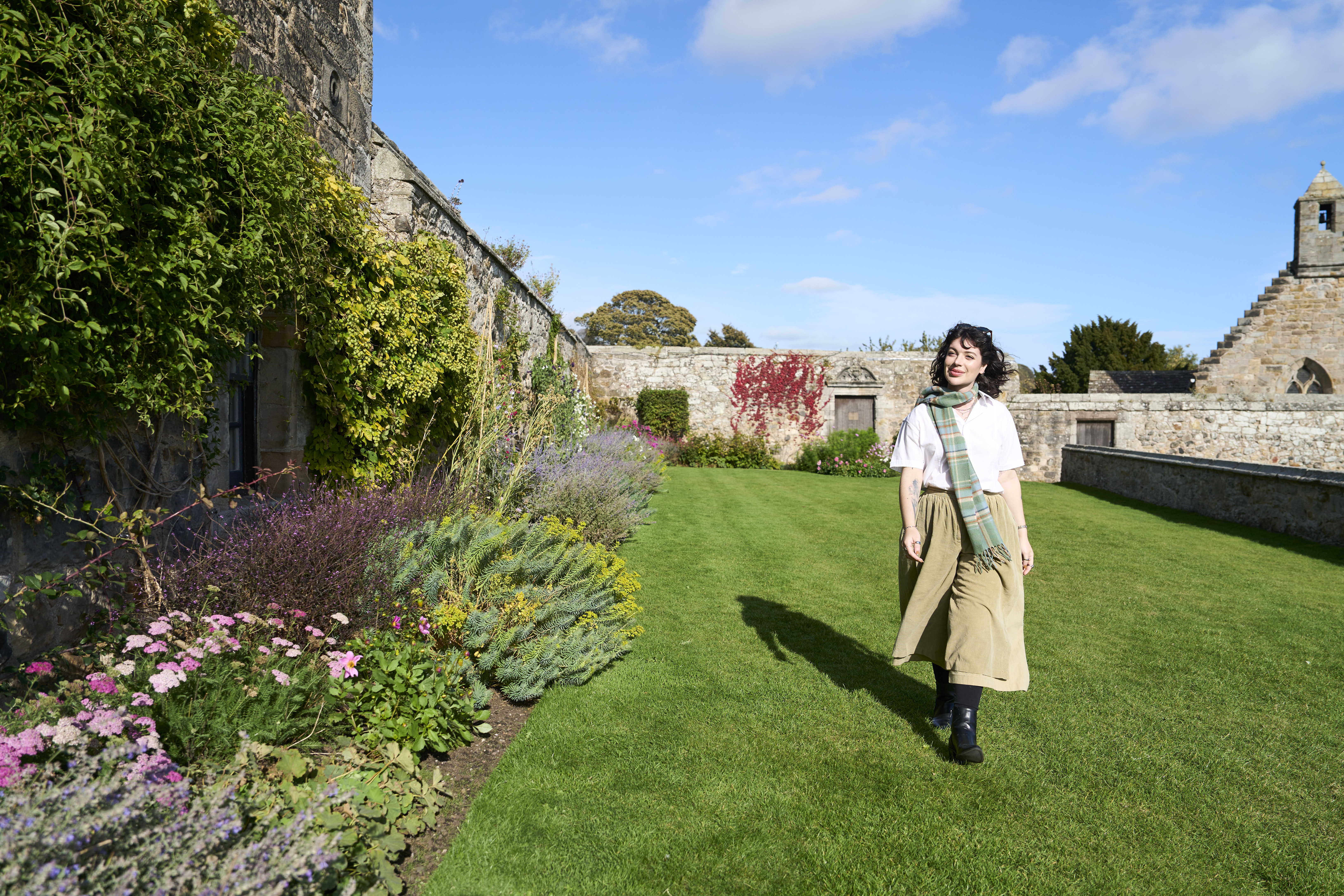 Frau geht im Garten neben einer Steinmauer, umgeben von Blumen und Pflanzen, unter blauem Himmel.