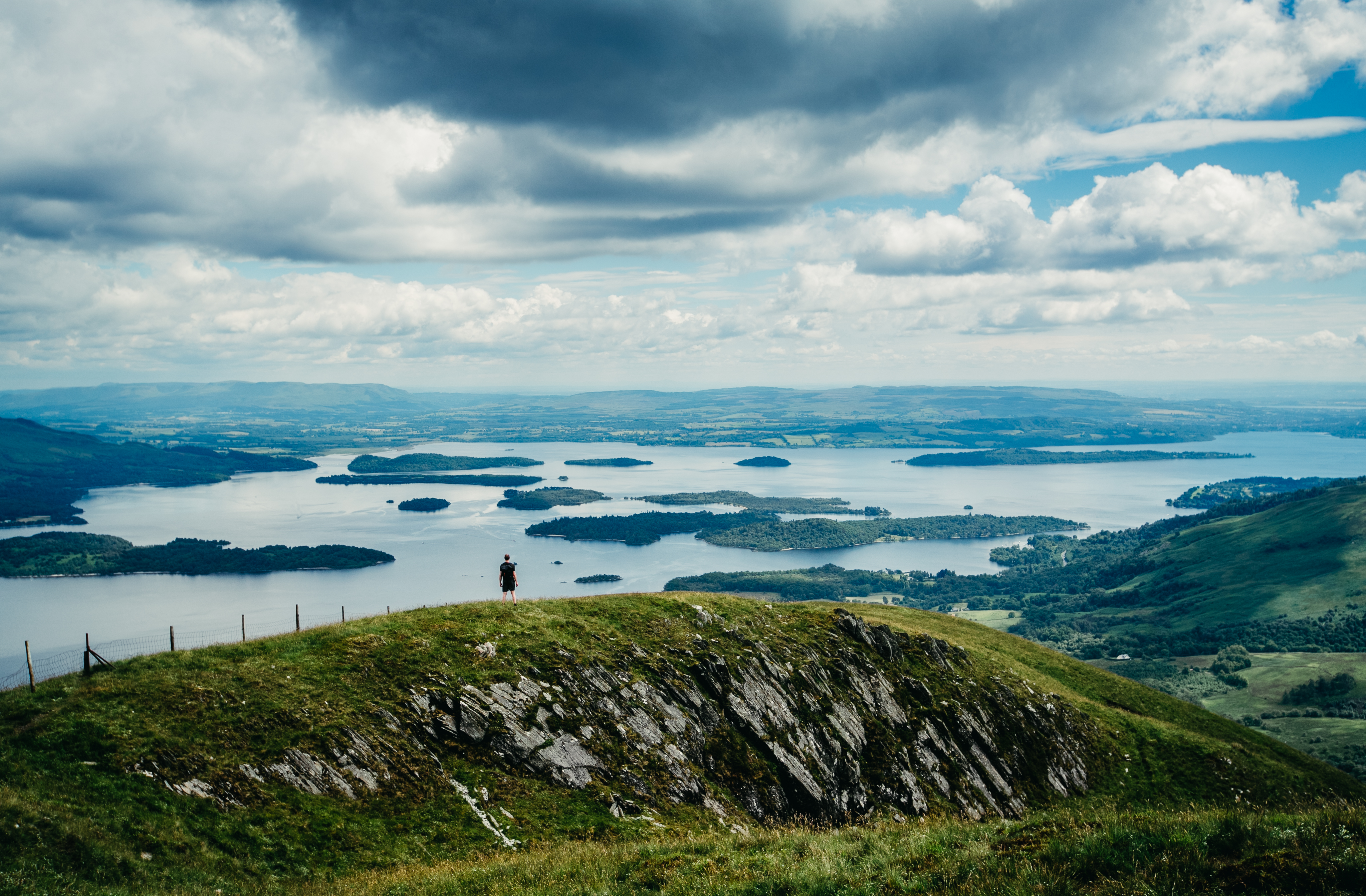 A wide panorama of a hiker standing atop a hill looking out over grassland and lakes.