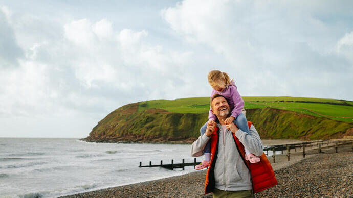 Person carrying a child on shoulders walking on a pebbled beach, with grassy hills and cloudy sky in the background.