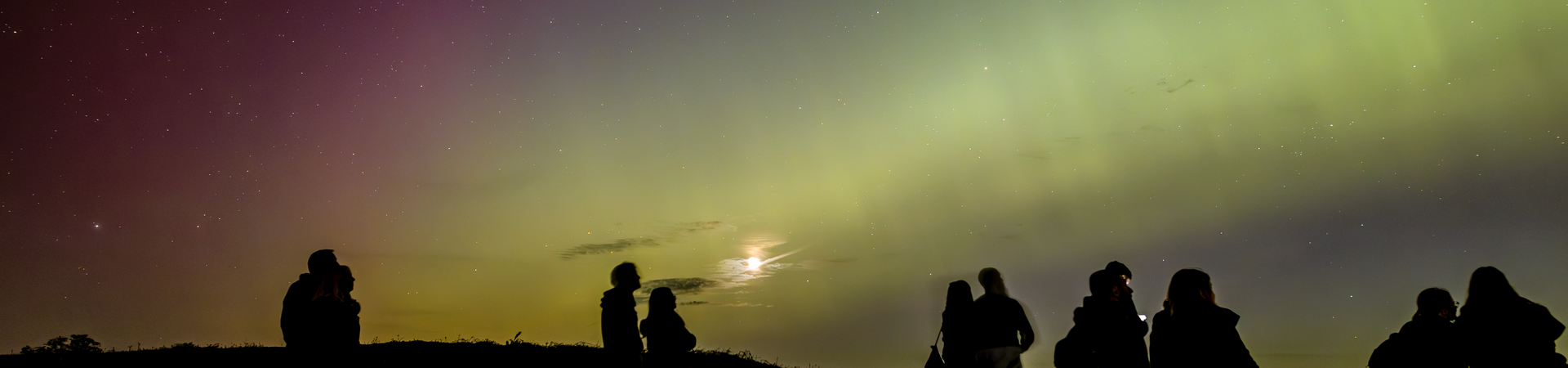 Silhouetted group of people watching the northern lights in a dark landscape under a colorful night sky.