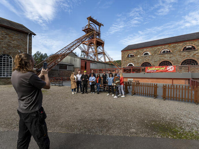 A group posing for a photo outside a historic mining site with stone buildings and metal mining tower under blue sky.