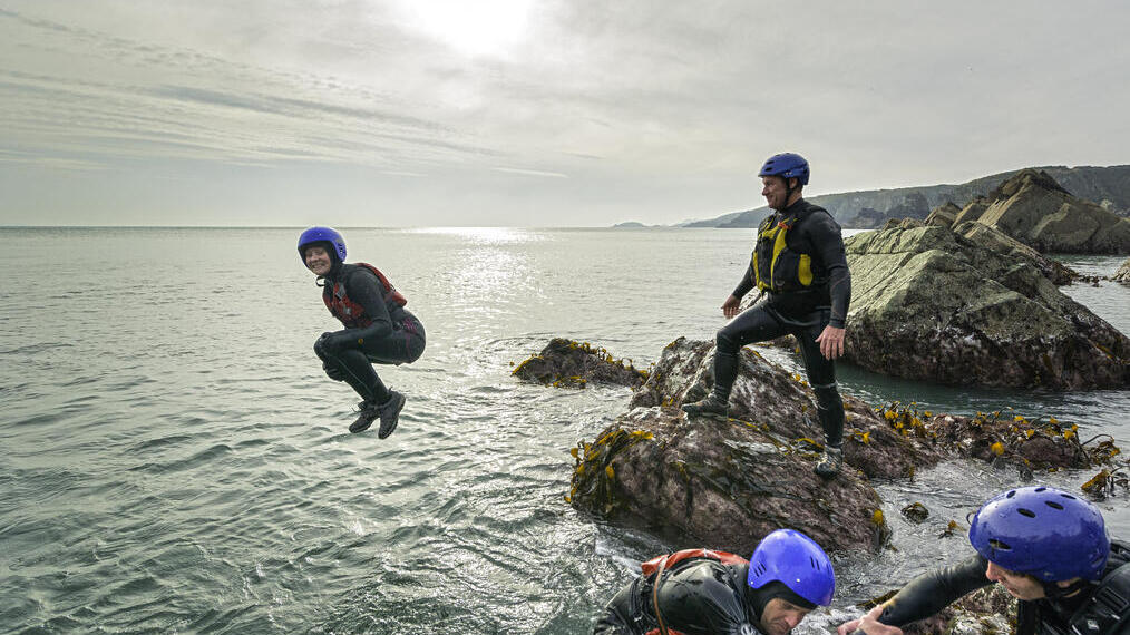 Grupo de personas con cascos y chalecos salvavidas en una actividad junto al mar; unas sobre rocas, una saltando al agua.