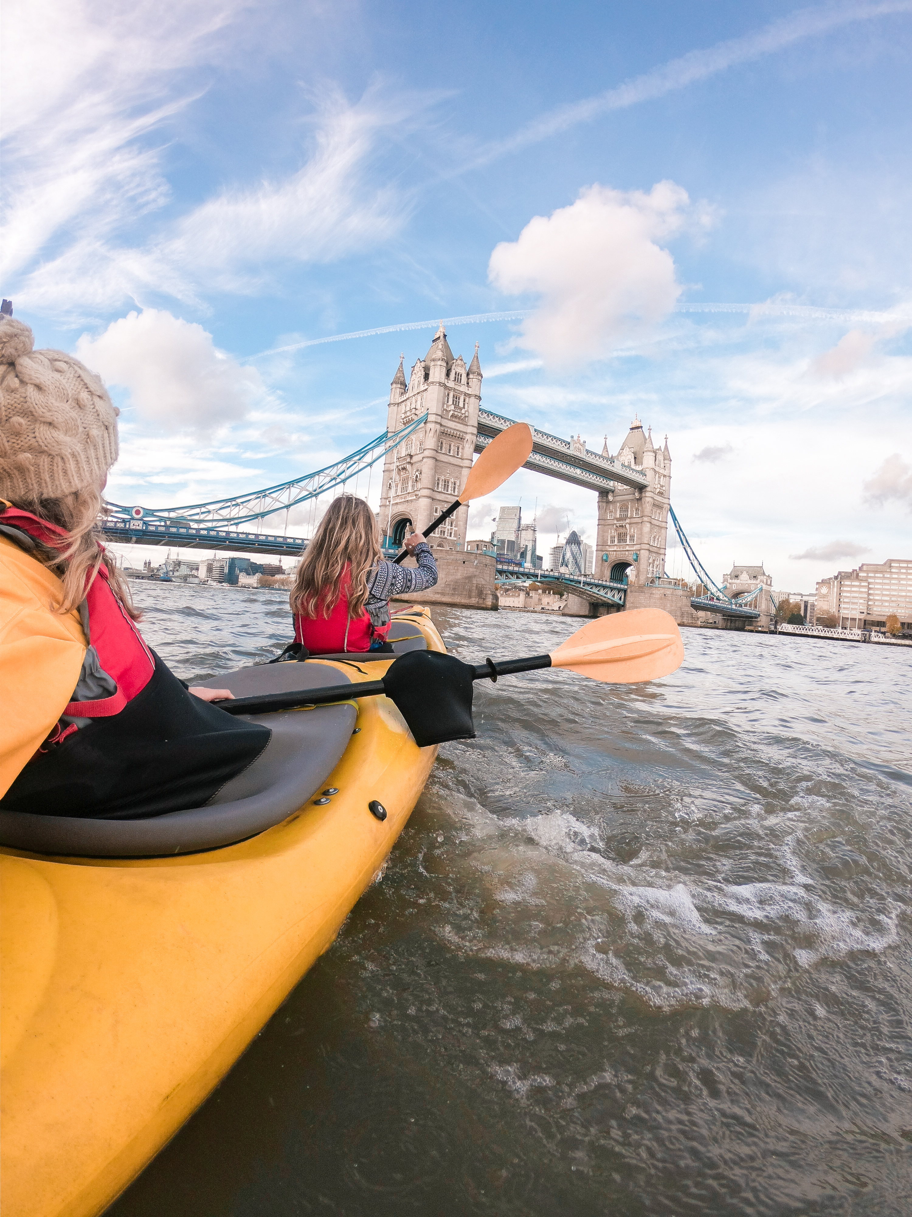 Two women paddling in a kayak on a river towards a bridge in a city
