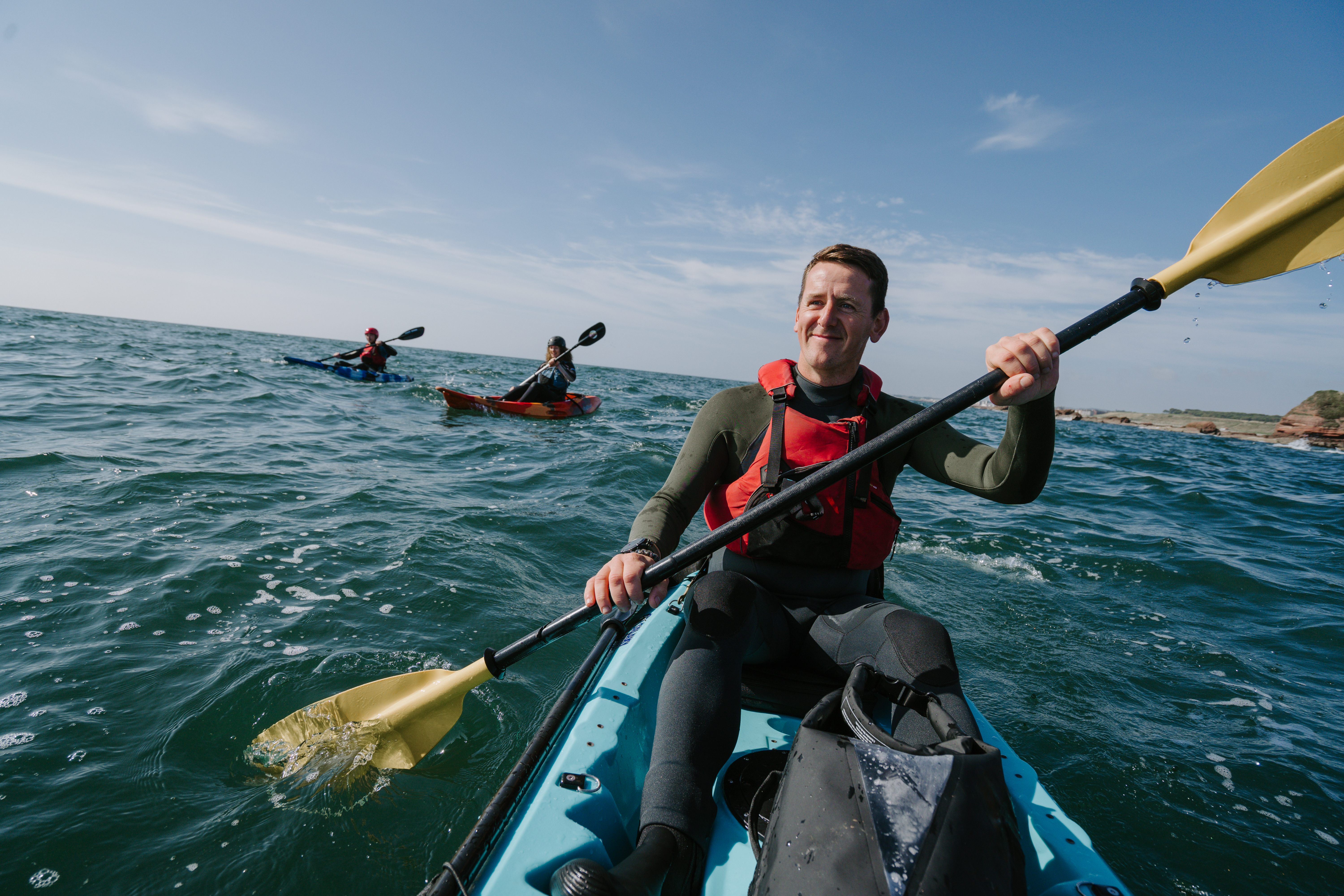 A man kayaking with two other people kayaking in the background.